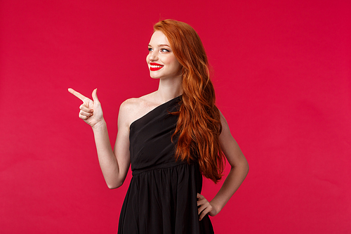 Portrait of sensual and feminine young redhead woman in elegant black dress, attend party, red carpet show, pointing finger left and looking satisfied with happy smile, beaming grin.