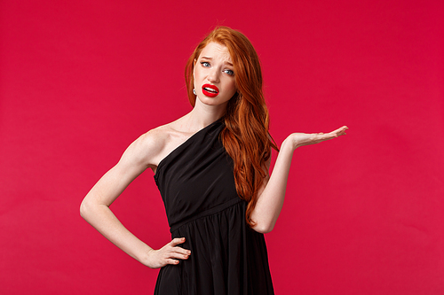 Whats your problem, so what. Arrogant and pissed-off snobbish sassy redhead woman in elegant black dress, shrugging with one head spread looking questioned and frustrated, red background.