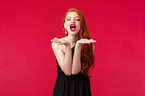 Portrait of passionate and sassy, seductive young beautiful woman with red hair in black dress, sending air kiss to camera, squinting flirty camera, seducing with her charm, stand red background.