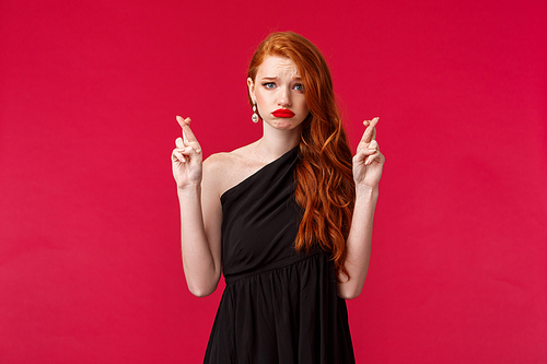 Portrait of hopeful worried caucasian female redhead in black luxurious dress, cross fingers good luck, want to win, making wish grimacing nervous, unconfident in own chances, red background.