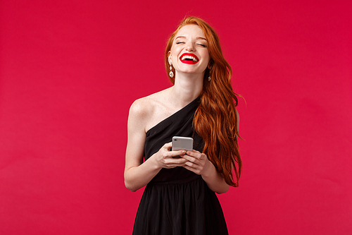 Technology and lifestyle concept. Gorgeous young redhead woman in black dress laughing over red background, holding smartphone, texting friend during prom discuss funny moment.