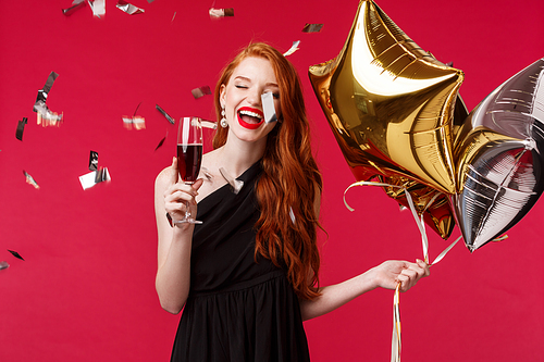 Portrait of carefree gorgeous feminine young redhead woman celebrating birthday, throw huge party, dancing and smiling with confetti flying air, hold balloons and glass wine or champagne, laughing.