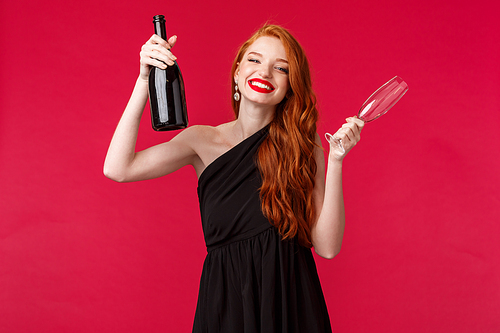Celebration, holidays and women concept. Portrait of gorgeous feminine young female with red hair, wear black stylish dress, raising bottle of champagne and glass, smiling camera.
