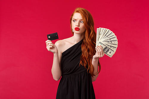Luxury, beauty and money concept. Portrait of indecisive and unsure young redhead woman pondering what to do with lots of cash dollars in her hands, looking at credit card hesitant.