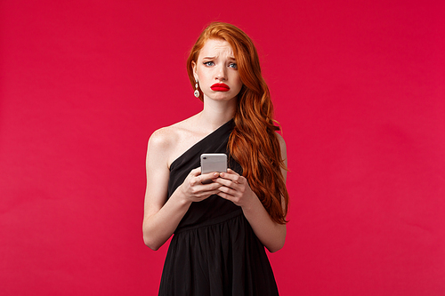 Portrait of gloomy upset gorgeous redhead girl in black dress, sobbing and grimacing unhappy, holding mobile phone, receive disappointing news via message, complaining, red background.