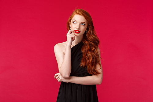 Portrait of thoughtful creative young redhead elegant woman with ginger hair in black dress, biting lip and looking up dreamy, thinking about solution, standing over red background.