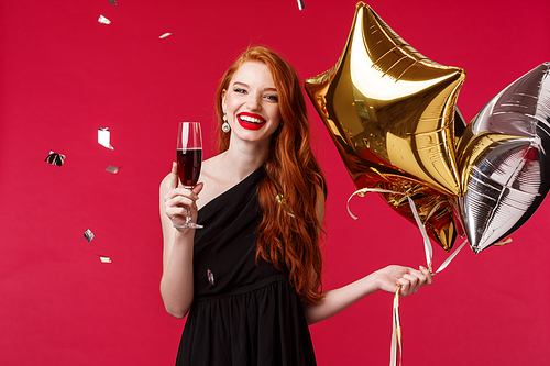 Celebration, holidays and party concept. Carefree gorgeous redhead woman having fun with friends throwing birthday, wear black dress, hold balloons and drink champagne with confetti in air.