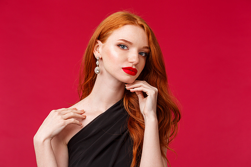 Close-up portrait of feminine good-looking seductive and sensual redhead woman in black dress, touching naked shoulder and gazing camera flirting, standing red background.