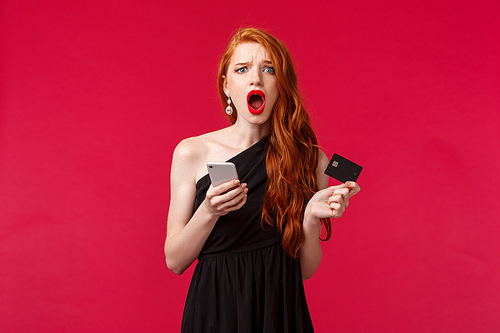 Portrait of complaining, disappointed gloomy clingy girlfriend with ginger long hair, feel unfair and jealous, holding credit card and smartphone, wanting buy something in internet, red background.