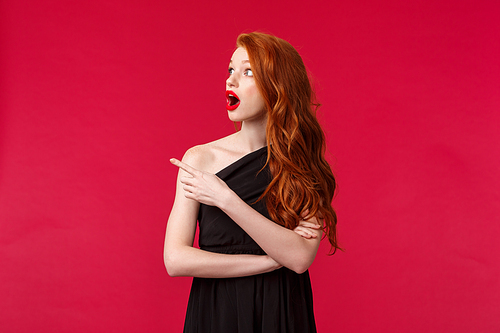 Celebration, events, fashion concept. Portrait of amazed and shocked redhead young pretty woman in elegant black evening dress, drop jaw, looking and pointing left astonished, red background.