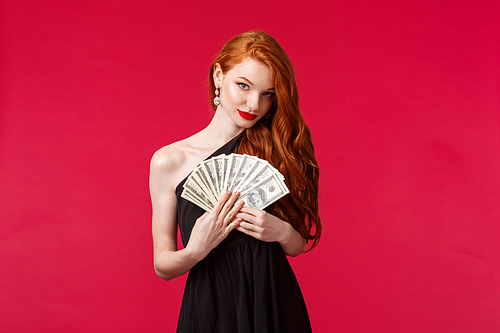 Luxury, beauty and money concept. Portrait of sensual good-looking, flirty young redhead woman in black stylish dress, hinting you can get this cash dollars if place good bet, smiling daring.