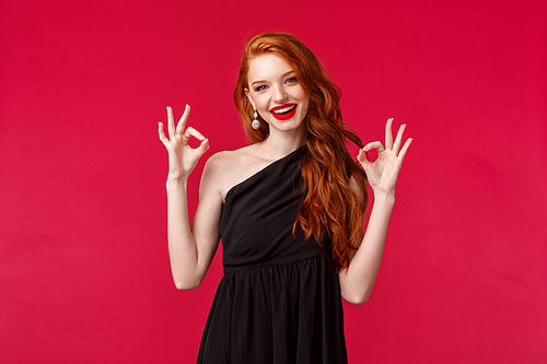 Fashion, luxury and beauty concept. Portrait of pleased good-looking sensual redhead woman in black dress and makeup, showing okay, excellent or guarantee sign, red background.