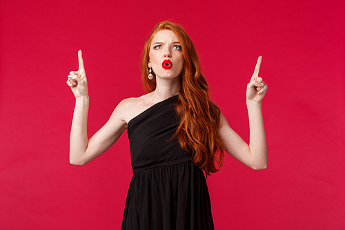 Celebration, events, fashion concept. Portrait of skeptical and displeased, young annoyed redhead woman in black dress, complaining grimacing unsatisfied, pointing fingers up bothered.