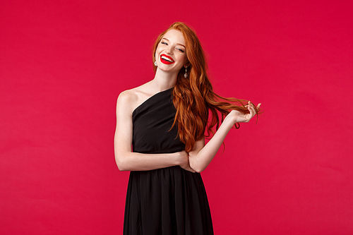 Elegance, fashion and woman concept. Portrait of confident sexy and stylish young redhead woman in red lipstick, evening makeup, wearing elegant black dress, laughing at formal event, red background.