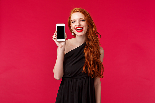 Portrait of adorable good-looking redhead woman with long ginger hair, wear black dress, makeup, showing mobile phone screen, recommend application, new filters app, smile camera, red background.