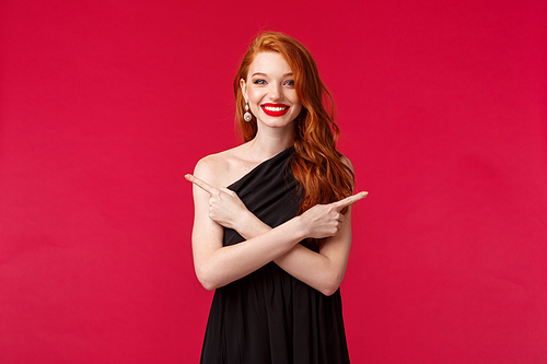 Fashion, luxury and beauty concept. Portrait of smiling redhead woman wearing makeup and black dress, pointing sideways at left and right, showing two variants, both choices good, red background.