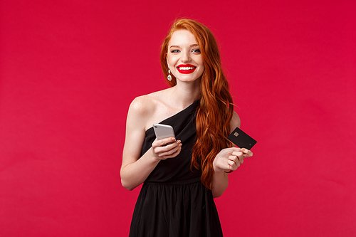 Portrait of charming young redhead girl in black dress, long ginger hair, holding credit card and smartphone, paying online purchase, order new outfit for party, stand red background.