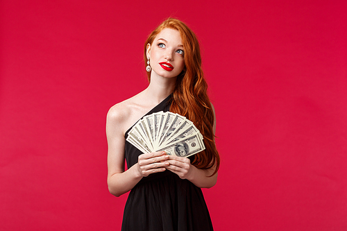 Luxury, beauty and money concept. Dreamy good-looking redhead woman with lots cash, become rich after receiving prize or reward, look up dreaming, thinking how waste income, red background.