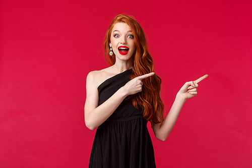 Celebration, events, fashion concept. Portrait of excited dreamy redhead woman in beautiful black dress, pointing fingers right and looking at camera astonished and impressed, red background.