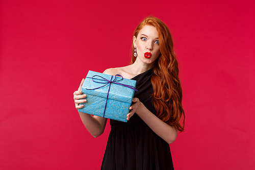 I wonder whats inside. Curious stunning young redhead woman in elegant black dress, shaking box with gift, folding lips amazed and intrigued look camera, receive present, red background.