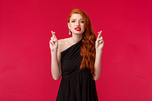 Portrait of worried hopeful redhead woman in black elegant evening dress, cross fingers for good luck, panicking and frowning nervously as pleading, await important news, stand red background.