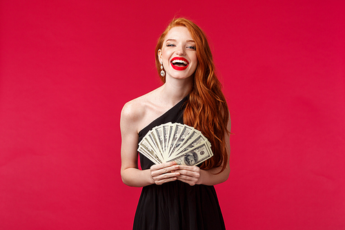 Luxury, beauty and money concept. Girl got expensive taste. Happy rich good-looking redhead woman in stylish black dress, laughing delighted, holding dollars, congratulate winner with prize.