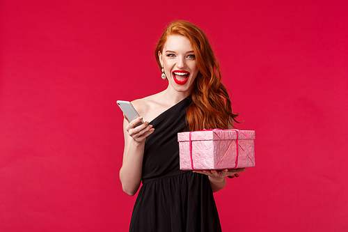 Portrait of lucky and sassy gorgeous redhead woman in red lipstick, black dress, showing happiness and joy, smiling daring look camera, hold gift box and smartphone, red background.