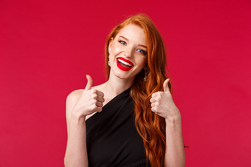 Close-up portrait of happy and satisfied redhead european woman recommend good place, wear black dress, party makeup, show thumbs-up in approval, smiling like performance, red background.