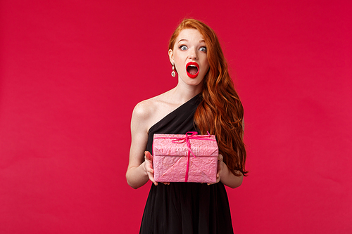 Celebration, holidays and women concept. Surprised b-day girl in elegant evening black dress, with ginger hair, drop jaw from excitement and happiness as receive awesome gift in pink box.