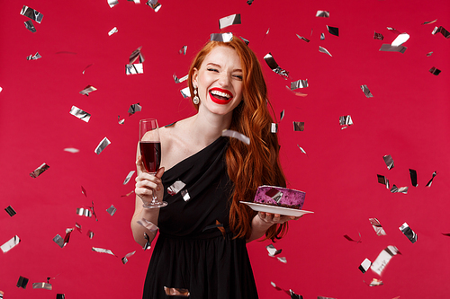 Celebration, holidays and party concept. Portrait of carefree beautiful young redhead woman having fun at birthday, celebrating laughing while confetti flying around, hold glass champagne and cake.