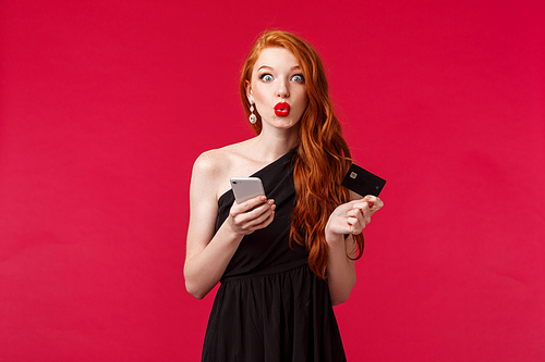 Portrait of excited good-looking female shopaholic in black stylish dress, holding smartphone and credit card, folding lips amazed, see special discount for birthday, stand red background.