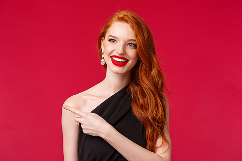 Close-up portrait of cheerful enjoyable young redhead girl in red lipstick, black dress, pointing and looking left with pleased intrigued expression, see something interesting, advertisement concept.