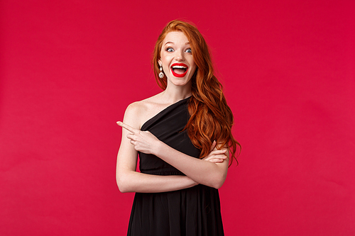 Elegance, fashion and woman concept. Portrait of excited happy redhead woman in elegant black slim dress, pointing finger left amazed smiling, hear great news, found perfect outfit for prom night.