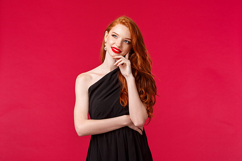 Fashion, luxury and beauty concept. Portrait of creative and dreamy attractive redhead female in black elegant dress, look up and smiling thinking, picturing nice thing in mind, red background.