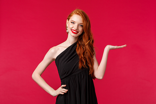 Portrait of elegant young redhead woman in luxurious black dress presenting something hold product on arm over red background, introduce company service, smile camera, show promo.