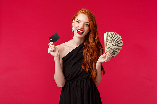 Luxury, beauty and money concept. Elegant good-looking redhead woman being rich, wear luxurious black dress, holding credit card and fan of dollars, brag her income, smiling carefree.