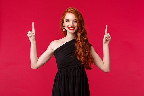 Celebration, events, fashion concept. Portrait of confident charming redhead female in elegant black dress, evening makeup, showing good store for buying prom outfit, pointing fingers up, smile.