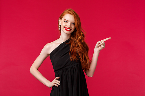 Fashion, luxury and beauty concept. Good-looking redhead woman with lipstick, evening makeup, showing way, click here, pointing finger right, smiling at camera wear elegant black dress.