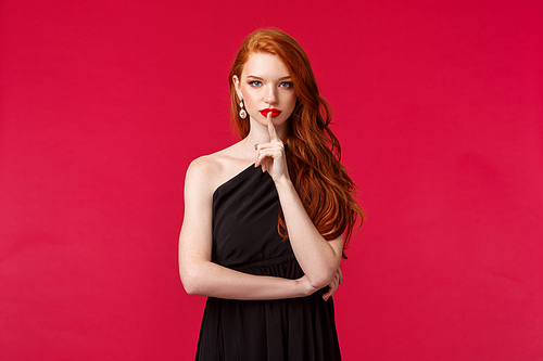 Fashion, luxury and beauty concept. Portrait of attractive sensual and seductive redhead woman in elegant black dress, show shush gesture, press finger to red lips and look camera serious.