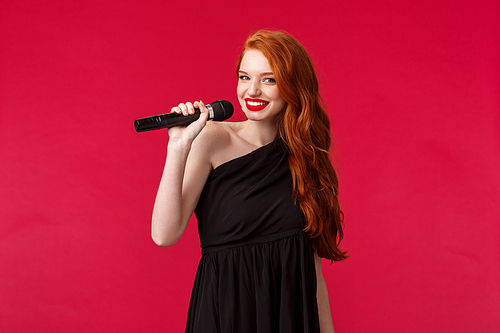 Portrait of fabulous young redhead woman with red lipstick, wear elegant evening black dress, hold microphone near lips, smiling camera as singing karaoke or performing, red background.