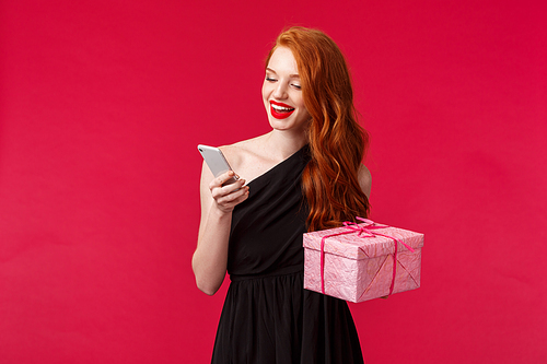 Portrait of cheerful elegant young redhead woman in black stylish dress, holding gift box and smartphone, texting friend thank you as receive present via mail, stand red background.