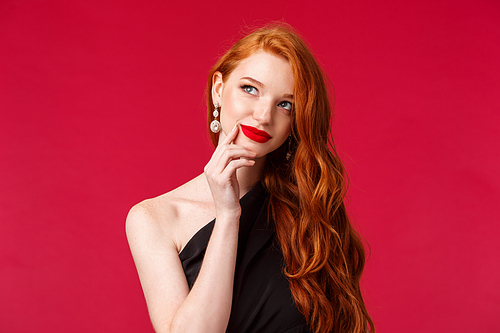 Close-up portrait of dreamy elegant and stylish redhead woman in black dress, red lipstick, making plan, thinking and smiling pleased of finding great idea, touch lip and look away pondering.