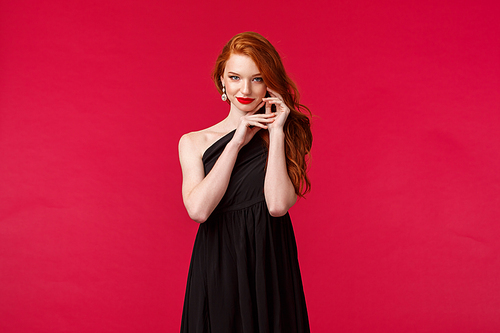 Elegance, fashion and woman concept. Portrait of seductive sensual redhead woman with red lipstick, wear evening stylish black dress over red background, look camera coquettish.