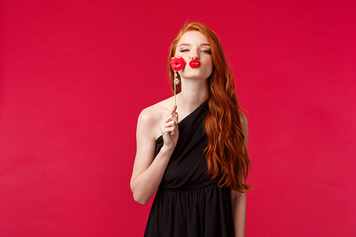 Portrait of gorgeous young redhead woman in evening black dress, feeling feminine and confident in herself, folding lips give kiss with red lipstick, holding paper lips stick, stand red background.