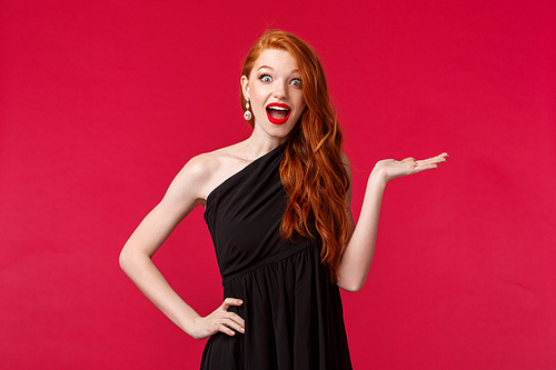Portrait of excited and happy young impressed ginger girl in slim black dress, look elegant, stare amazed at camera fascinated as holding something in arm over red background.