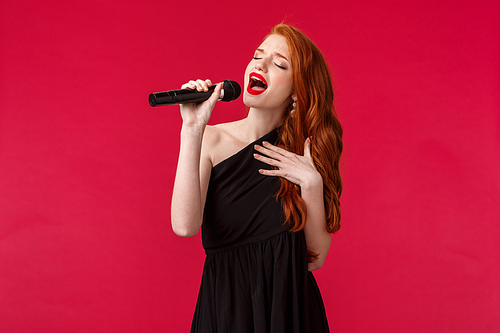 Close-up portrait of passionate beautiful female singer perform songs, wearing black evening dress, close eyes and showing her feelings through music, hold microphone, attend karaoke on girls night.