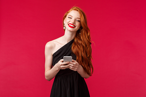 Portrait of gorgeous smiling woman with ginger hair in elegant black prom dress, holding smartphone, messaging, using application, want take picture, laughing and looking camera, red background.