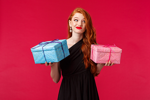 Celebration, holidays and women concept. Portrait of indecisive good-looking redhead woman in black dress, holding two gifts in hands, look away and smirk indecisive, dont know what give.