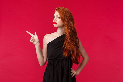 Portrait of sad and pity cute redhead woman in elegant black dress, sympathizing showing empathy, grimacing distressed and lonely, pointing looking left with regret, red background.