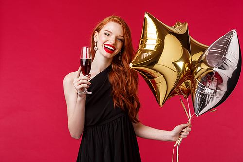 Portrait of happy carefree, gorgeous redhead woman in elegant black dress partying, saying toast, cheers holding glass of champagne or wine, smiling happy with balloons in hand, red background.